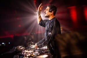A DJ wearing headphones performs energetically with one hand raised, surrounded by sound equipment and colorful lights.