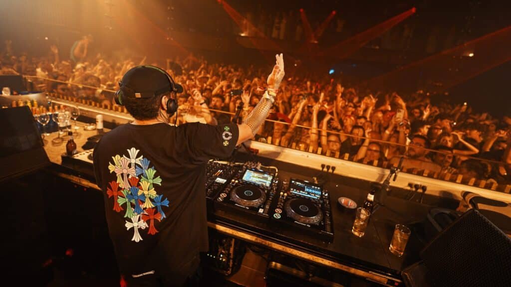 A DJ wearing headphones and a floral shirt plays music for a large, energetic crowd at a club, with vibrant lighting.