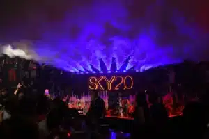 A crowded nightclub with vibrant blue and red lighting, featuring a neon "SKY20" sign and smoke effects above the bar.