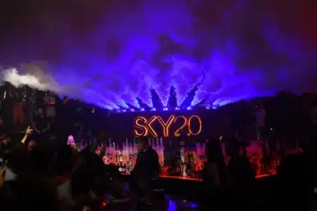 A crowded nightclub with vibrant blue and red lighting, featuring a neon "SKY20" sign and smoke effects above the bar.