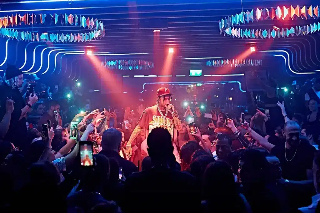 A performer on stage is surrounded by a crowd taking photos and videos with their phones in a dimly lit, colorful nightclub.