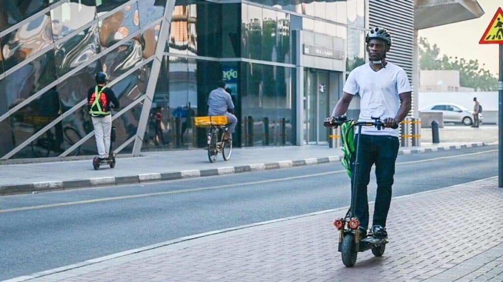 A man wearing a helmet rides an electric scooter on a city sidewalk, with a biker and another scooter in the background.