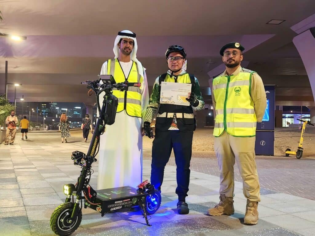 Three men in reflective vests stand together; one holds a certificate and an electric scooter, under a city bridge at night.