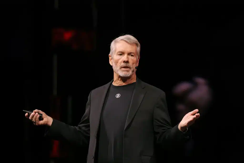 A man with gray hair and a beard speaks on stage, wearing a black suit and holding a microphone.