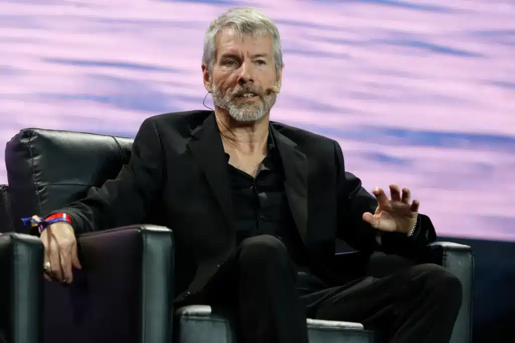 Michael Saylor speaking into a microphone about Satoshi's Gift while seated on a stage, wearing a black suit.