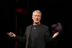 A man with gray hair and a beard speaks on stage, wearing a black suit and holding a microphone.