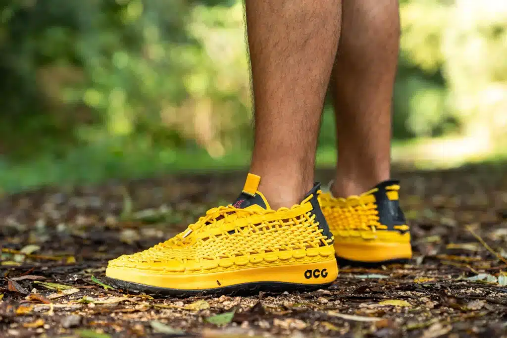 Person wearing bright yellow woven sneakers with black accents, standing on a leafy ground.