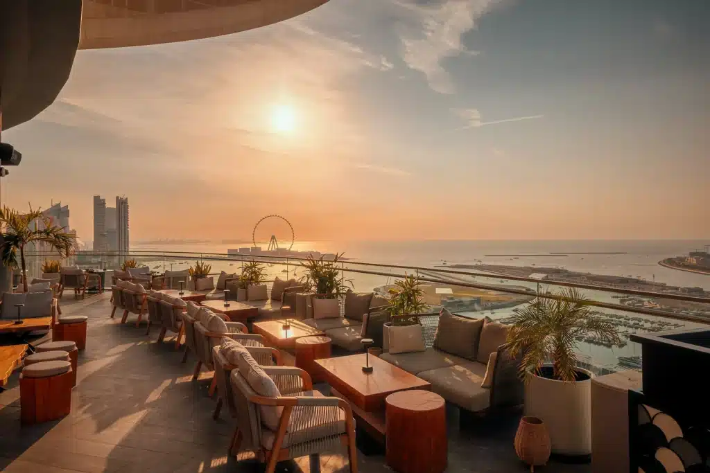 A rooftop lounge with comfortable seating overlooks a coastal cityscape and a Ferris wheel at sunset.