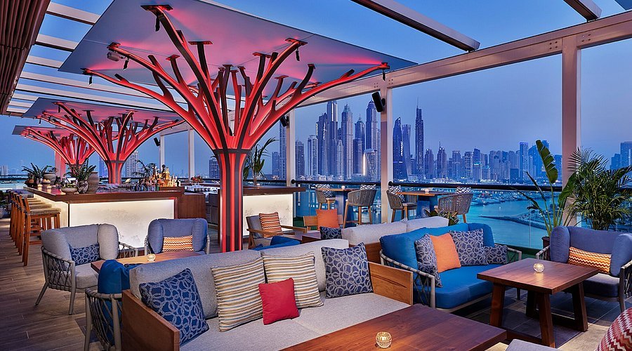 A stylish rooftop bar with modern seating, red-lit tree structures, and a city skyline view at dusk.
