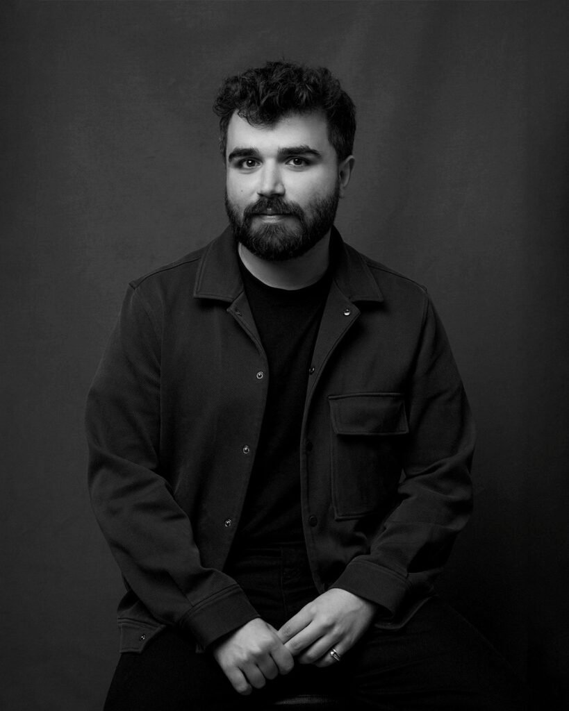 A man with a beard and curly hair sits against a plain dark background, wearing a dark jacket and shirt.