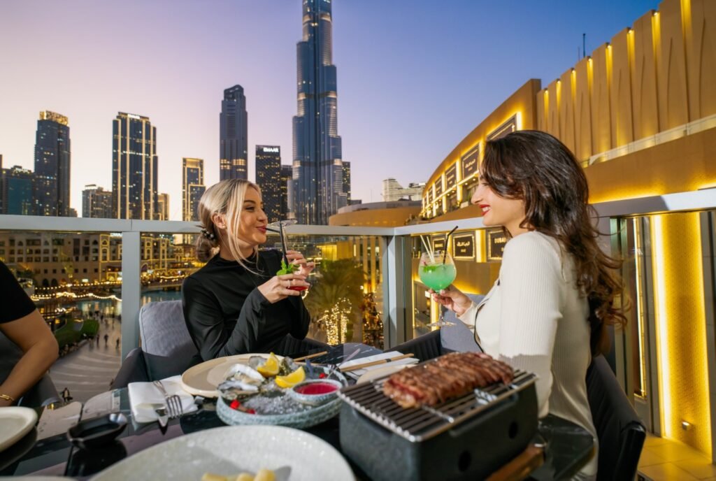 Two women enjoying drinks at a rooftop restaurant with skyscrapers in the background at sunset.