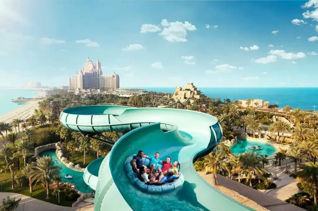A group of people on a raft sliding down a twisty waterslide at Aquaventure Waterpark, with a view of palm trees and a large resort in the background.
