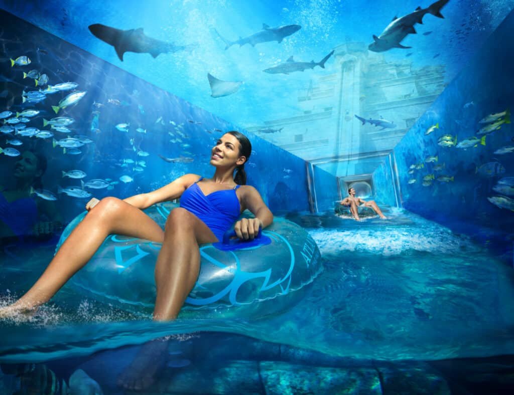 A woman in a blue swimsuit floats in a tube through an underwater tunnel in Aquaventure Waterpark surrounded by various fish and sharks.