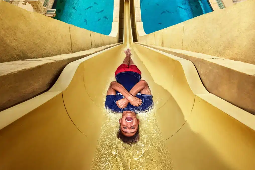 Person sliding down a water slide, wearing a black shirt and red shorts, with an expression of excitement.