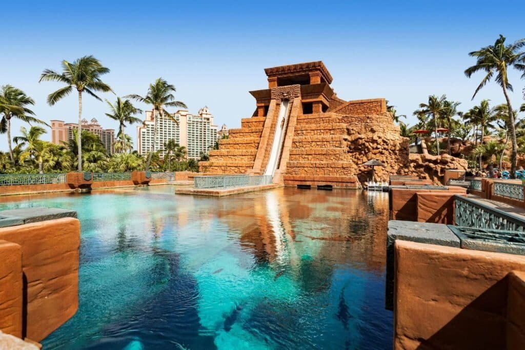 A pool with clear blue water reflecting a Mayan pyramid-style water slide, surrounded by palm trees.