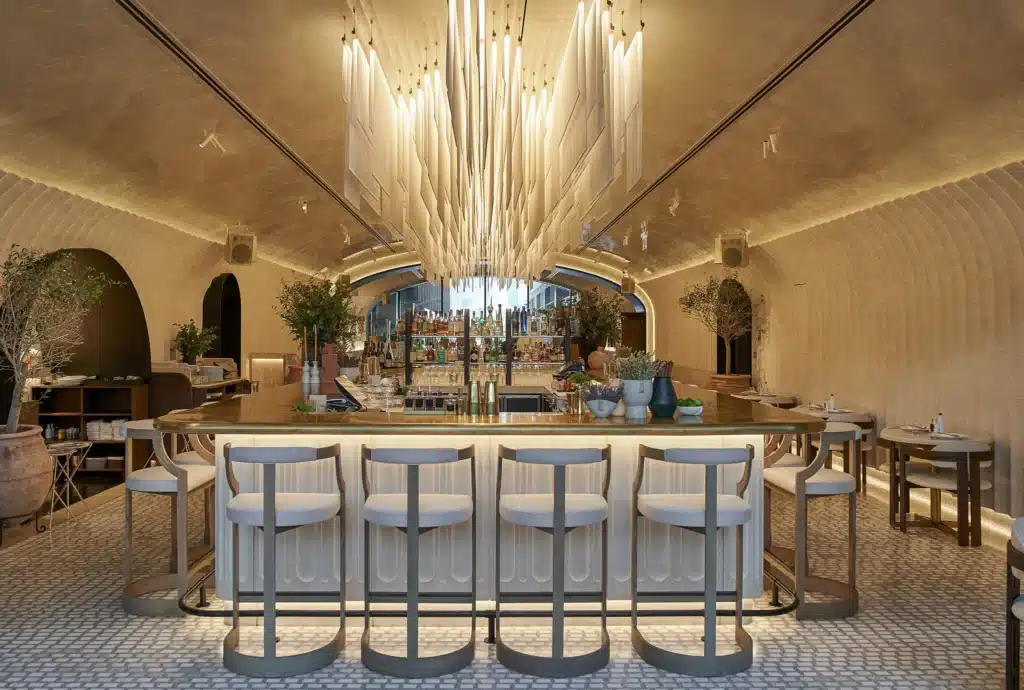 Dubai Best Restaurant Deals: Stylish bar interior with a large central chandelier, curved seating around the bar, and plants in the background.