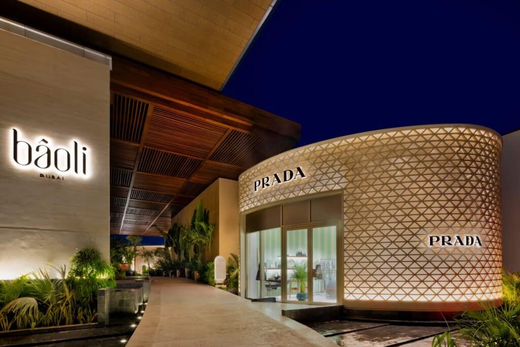 Exterior view of Bâoli Dubai and Prada stores at night, with modern architecture and illuminated signs.