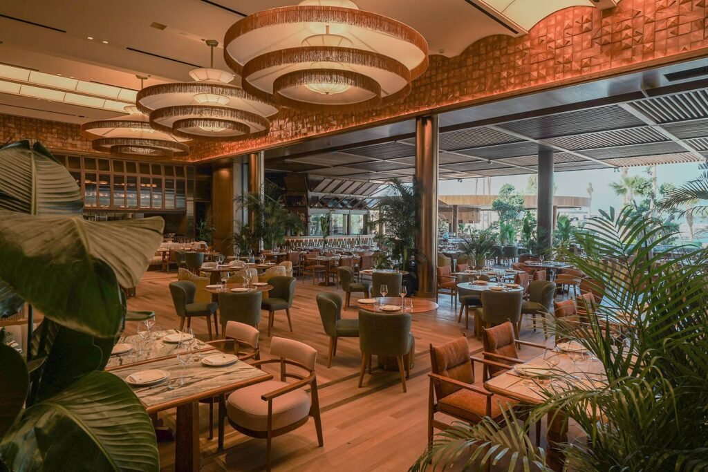 Elegant restaurant interior with warm lighting, wooden flooring, and tables set with glassware, surrounded by lush green plants.
