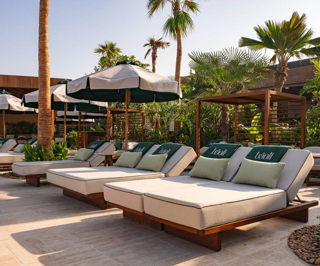 Outdoor Bâoli Dubai lounge area with cushioned wooden sunbeds, green umbrellas, and lush palm trees in a tropical setting.
