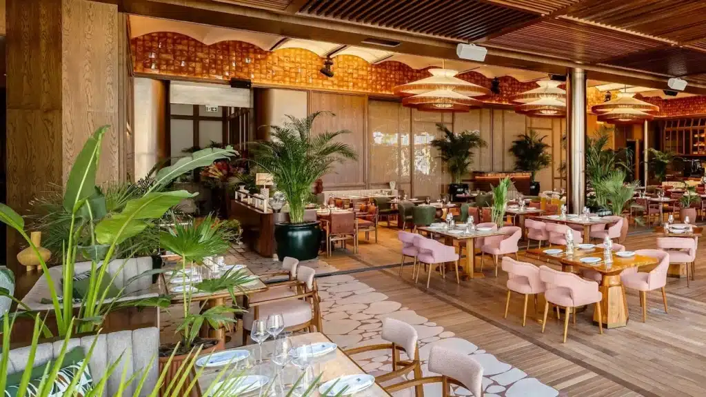 A spacious, elegant restaurant interior with wooden flooring, pink cushioned chairs, and tables set for dining, surrounded by large plants.
