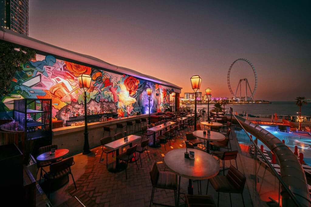Rooftop bar with colorful graffiti-covered walls, ambient lights, and a view of a Ferris wheel at sunset near the water.