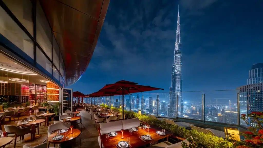 A rooftop restaurant with tables set for dining, overlooking a vibrant city skyline with the Burj Khalifa prominently visible at night.