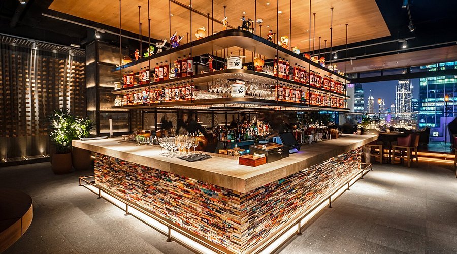Modern bar with illuminated shelves stocked with liquor bottles, a central counter, and a cityscape visible through large windows.
