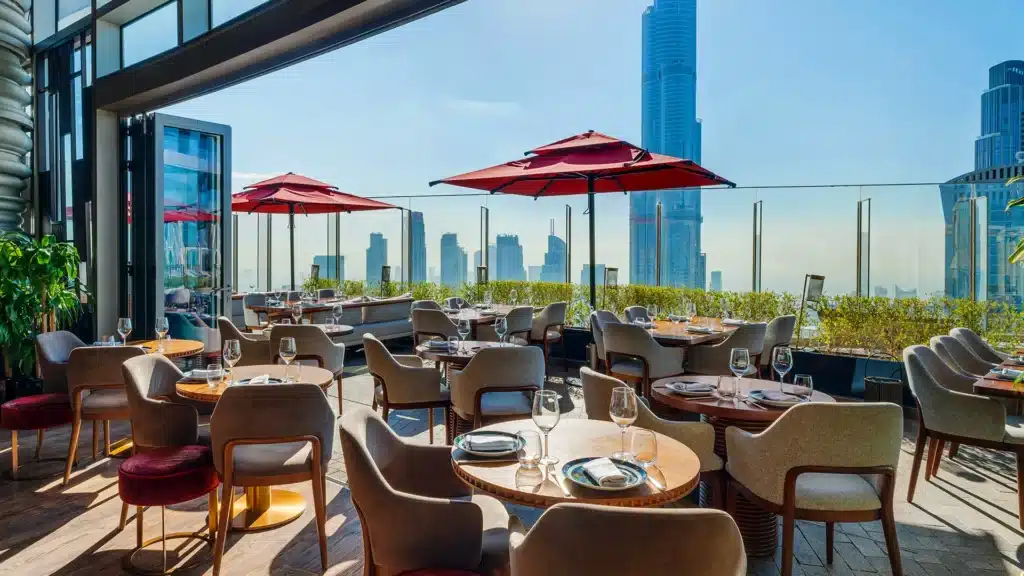 Outdoor restaurant seating with tables set for dining, red umbrellas, and a city skyline view in the background.