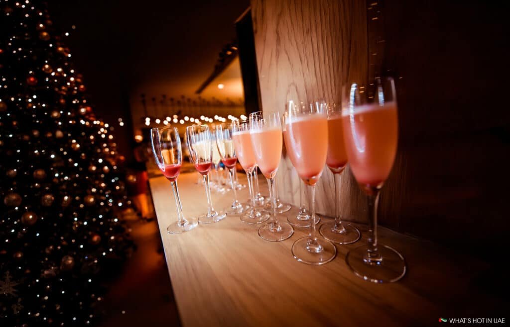 Glasses of pink cocktails lined up on a wooden surface, with a decorated Christmas tree in the background.