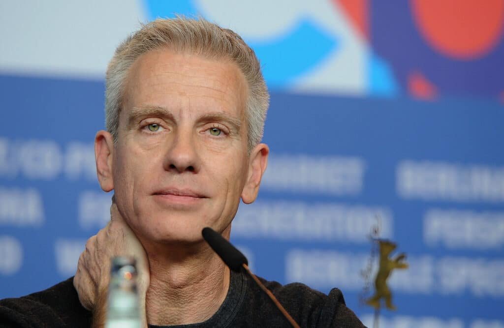 Chris Sanders with short gray hair attends a press event, with a blue backdrop featuring blurred Lilo & Stitch 2 text and a bear logo.