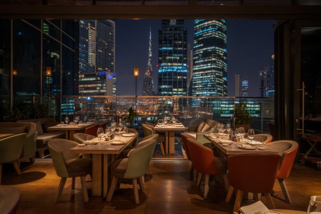 Elegant restaurant interior with tables set for dining, large windows showing a city skyline illuminated at night.