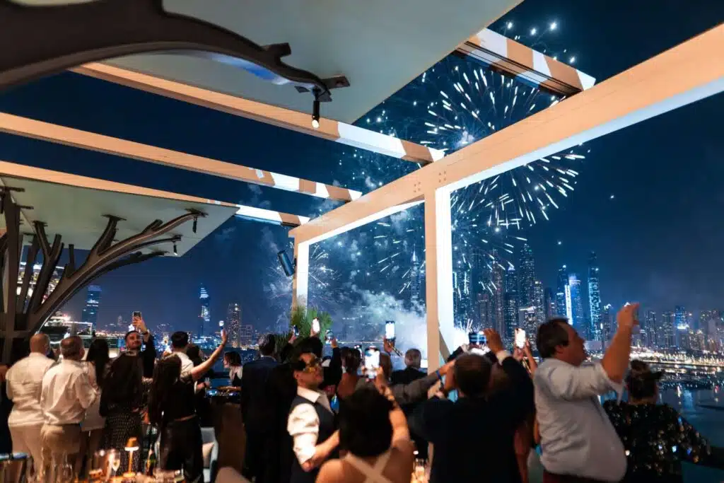 A group of people on a terrace watch fireworks over an illuminated city skyline at night.