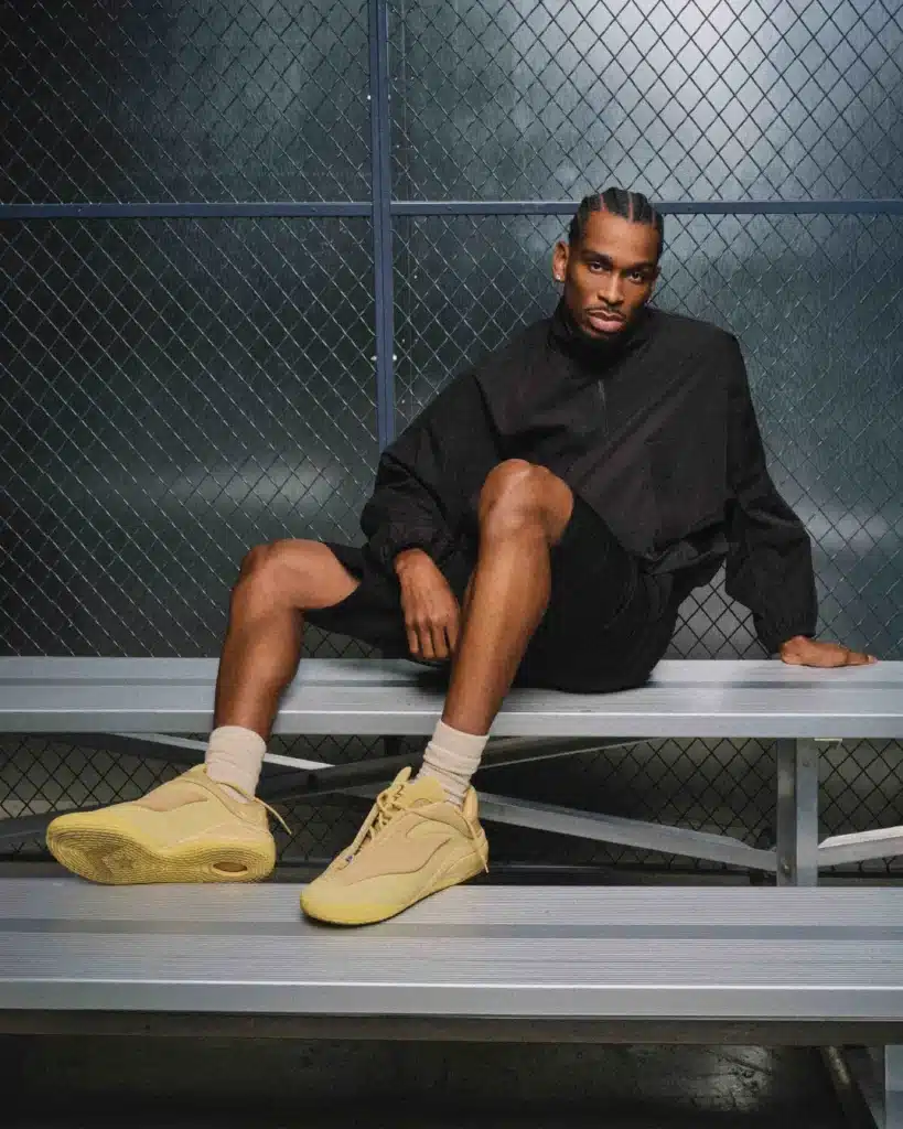 A person in a black outfit with braided hair is sitting on metal bleachers, wearing yellow Converse SHAI 001 sneakers.