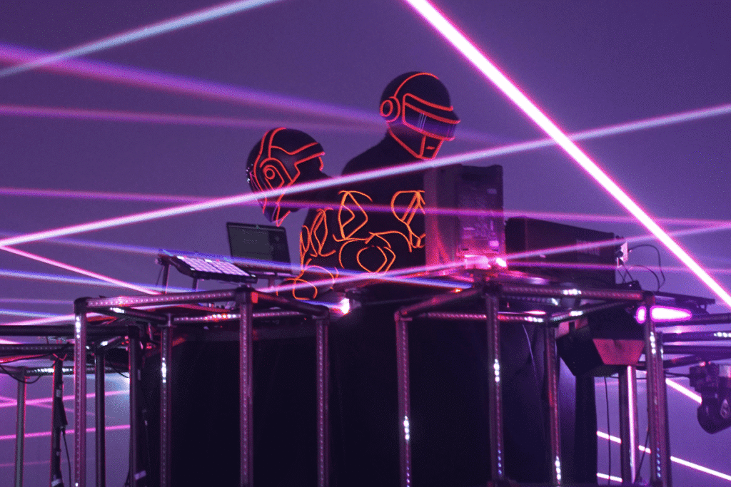 Daft Punk in illuminated helmets and suits stand behind DJ equipment with vibrant laser lights cutting through the scene.