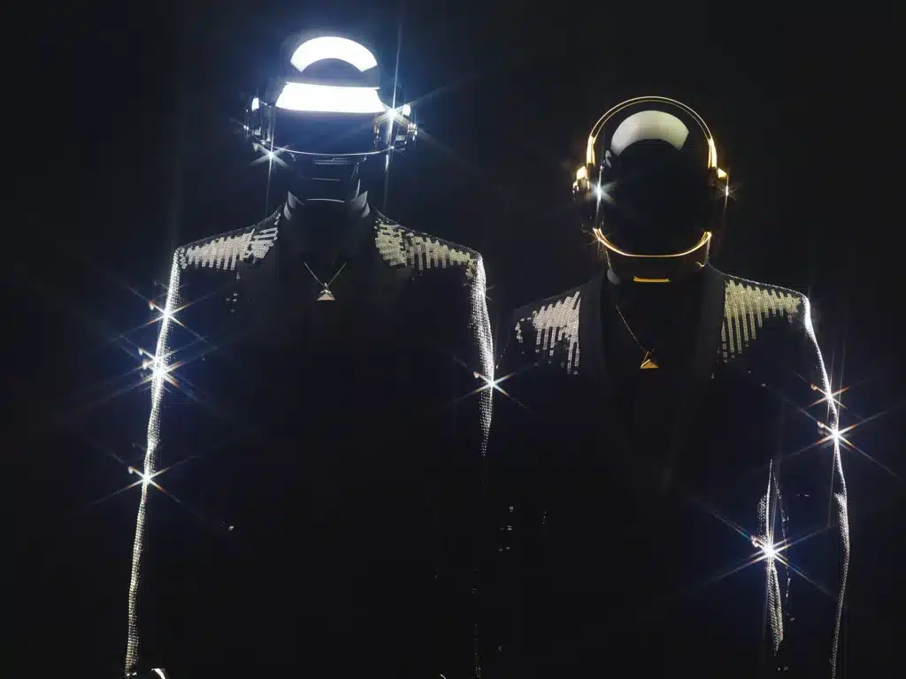 Two individuals wearing shiny, sequined suits and futuristic helmets with illuminated accents stand side by side in a dark setting.