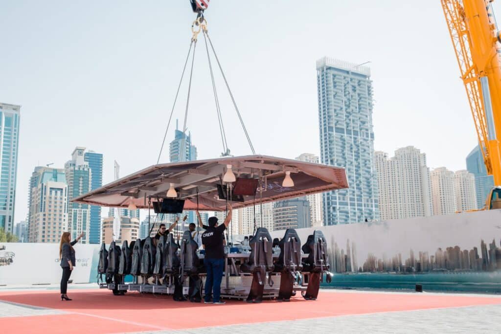 A platform with dining seats is suspended by a crane against a city skyline background.