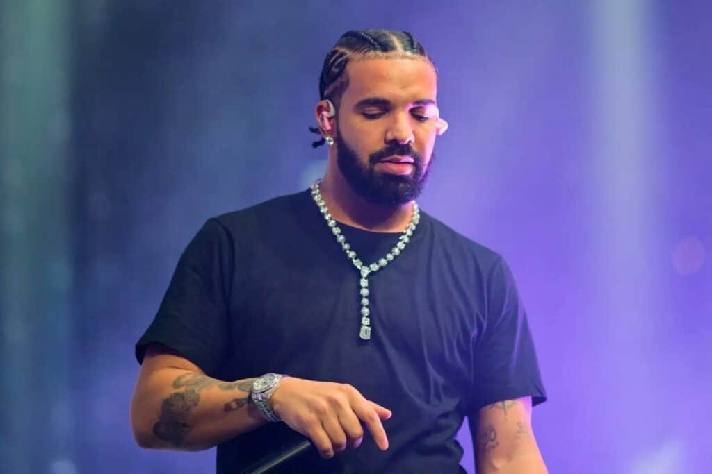 Drake with braided hair holds a microphone on stage, wearing a black shirt and a diamond necklace, under purple lighting.