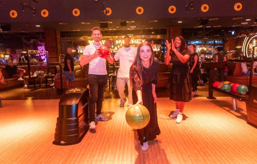 Four people at a bowling alley, one woman in front about to bowl a green ball, with others smiling and clapping behind her.