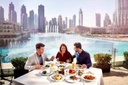 Three people enjoy a meal at an outdoor table overlooking a city skyline and large fountain.