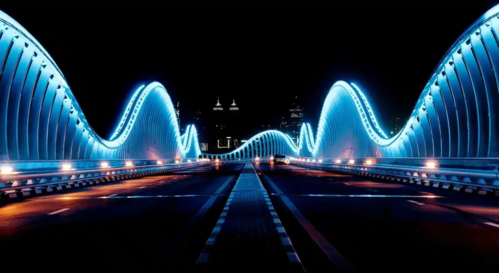 A futuristic bridge at night with wavy, illuminated railings in blue light, leading toward a city skyline in the distance.