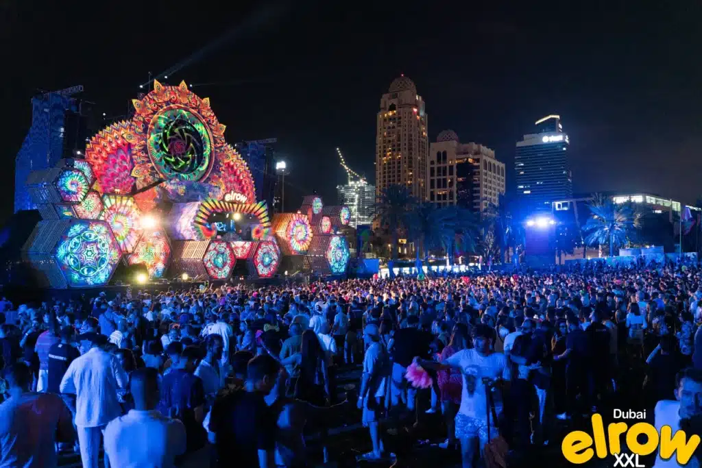 A large crowd enjoys a vibrant, colorful stage with intricate light displays at an outdoor nighttime Elrow Dubai music festival in Dubai.