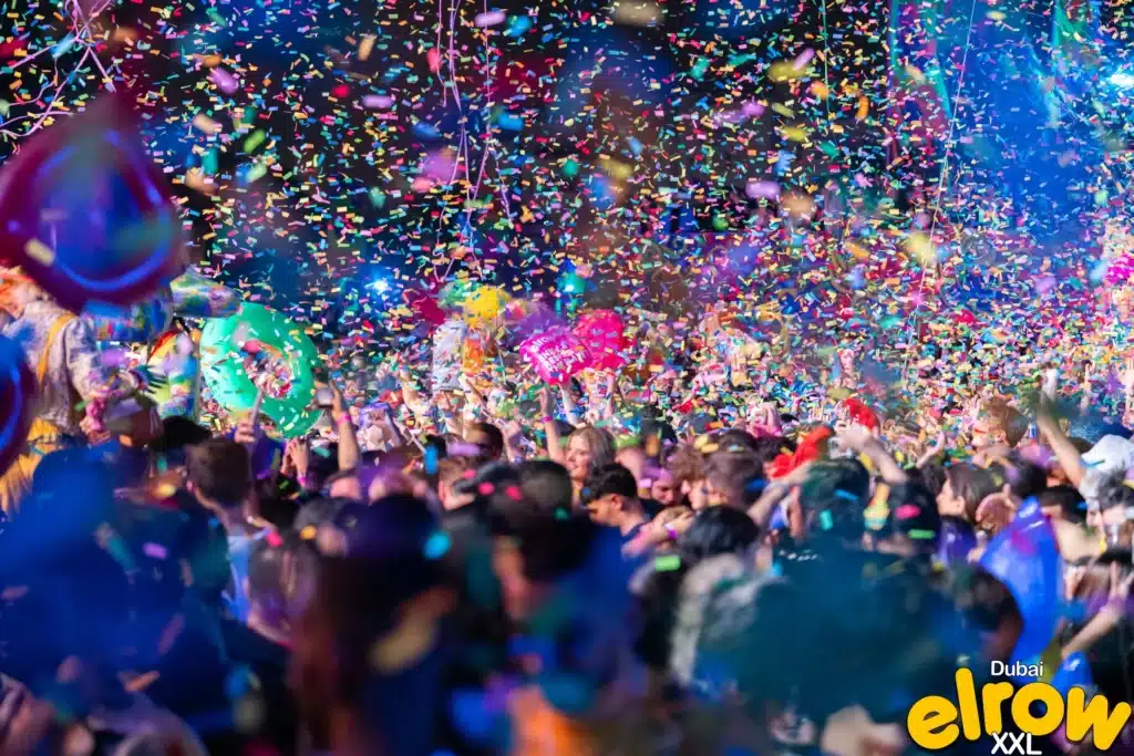 A vibrant crowd at Elrow Dubai with colourful confetti falling and people holding inflatable decorations.