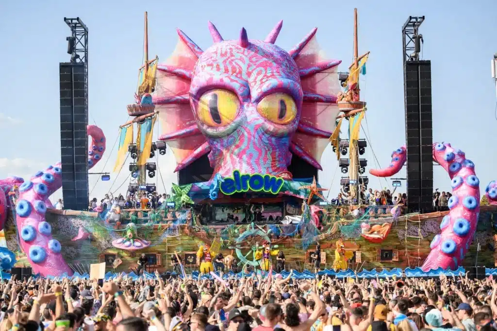 A colorful festival stage with a large, vibrant sea creature design and "elrow" signage, surrounded by a lively crowd.
