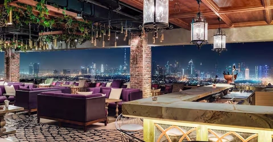 Rooftop lounge with purple sofas and lanterns, overlooking a city's illuminated skyline at night.