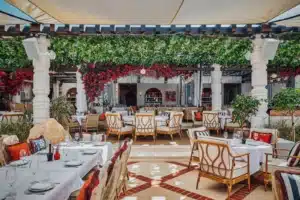 Outdoor restaurant with white tablecloths, wicker chairs, and leafy green and red floral decor under a shaded canopy.