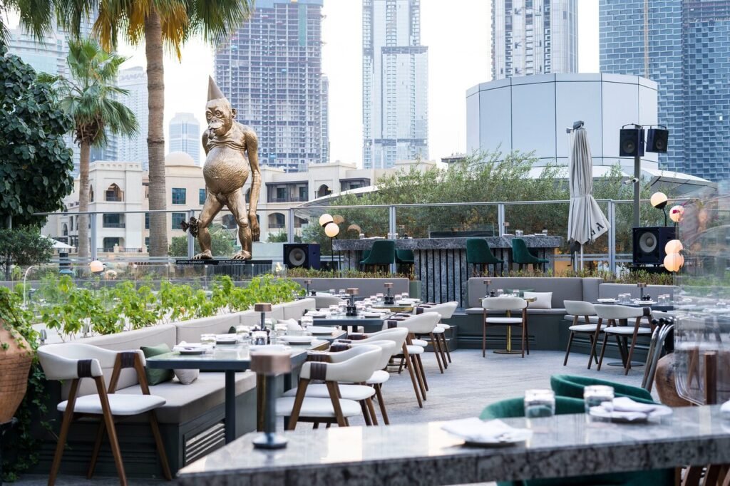 An outdoor GĀL Dubai restaurant with neatly set tables and chairs, featuring a large, unique statue and a city skyline in the background.