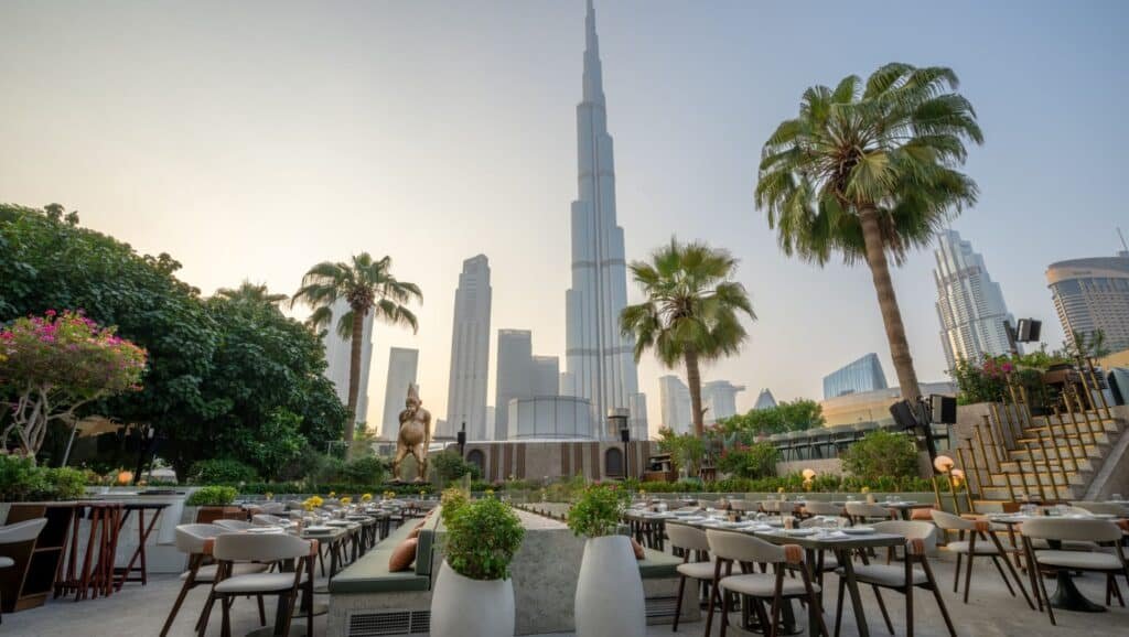 Outdoor restaurant with neatly arranged tables, lush greenery, and a clear view of a tall skyscraper in the background.