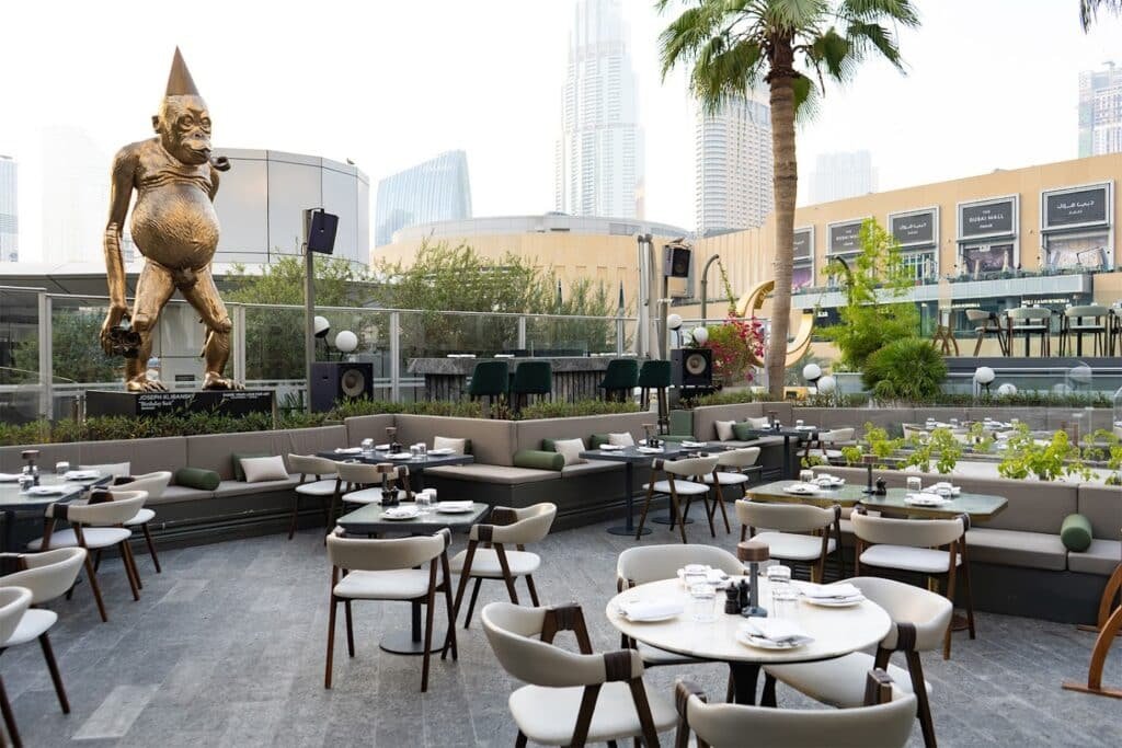 Outdoor GĀL Dubai dining area with empty tables and chairs, featuring a large sculpture of a creature with a pinecone head in the background.