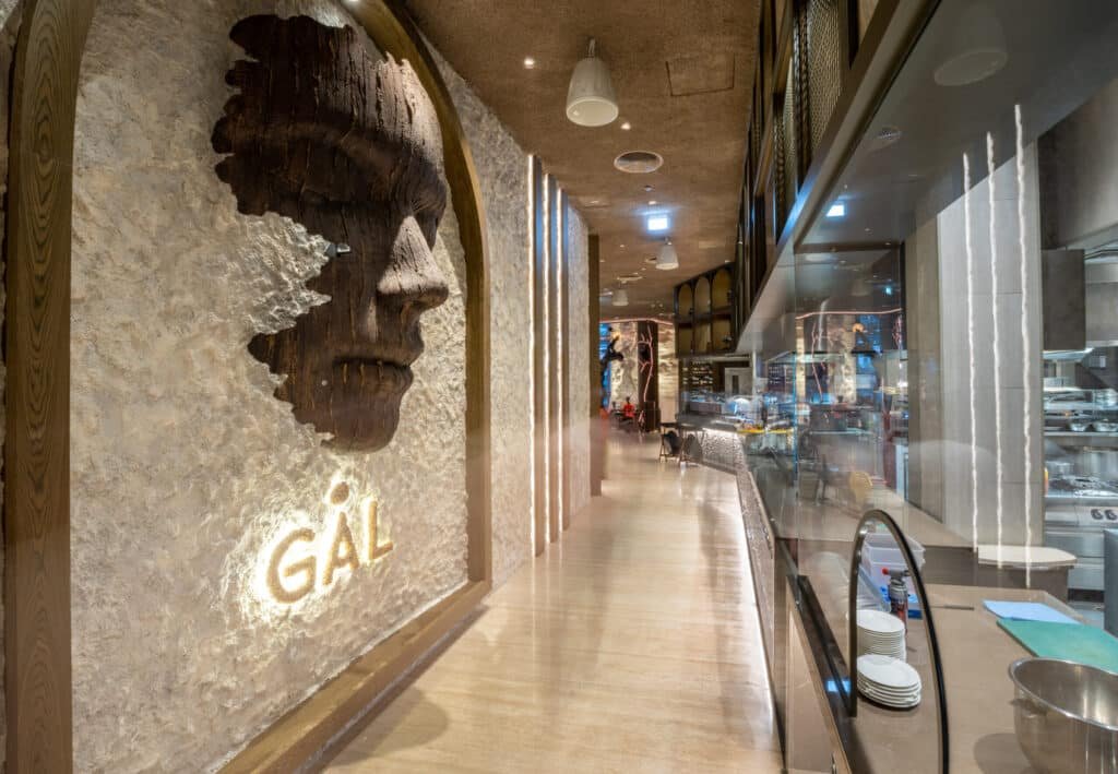 Interior of a restaurant with a large, textured wall sculpture of a face and the word "GÁL" illuminated beside it.