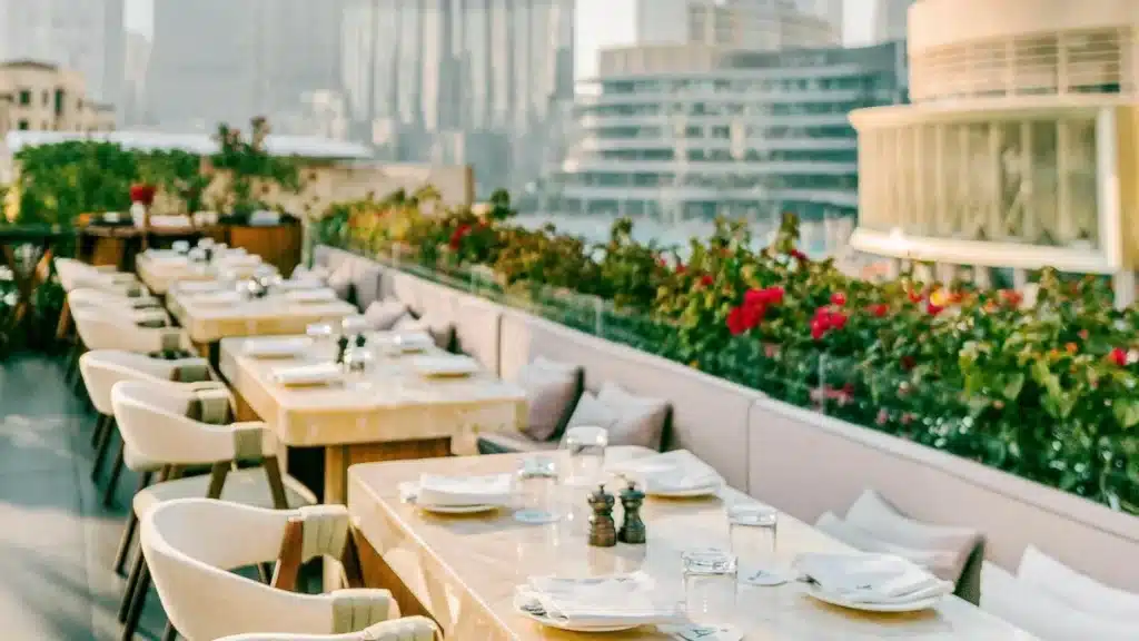 Outdoor restaurant terrace with neatly set tables, cushioned chairs, and a cityscape backdrop, bordered by vibrant flowers.